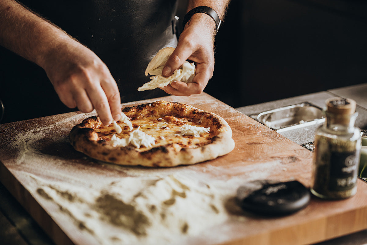 Frisch gebackene Pizza wird auf einem Holzbrett mit Käse belegt
