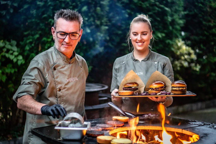 Grillmeister der MOESTA Grillschule grillt Burger an der Feuerplatte, daneben eine Servicekraft mit gefülltem Tablett