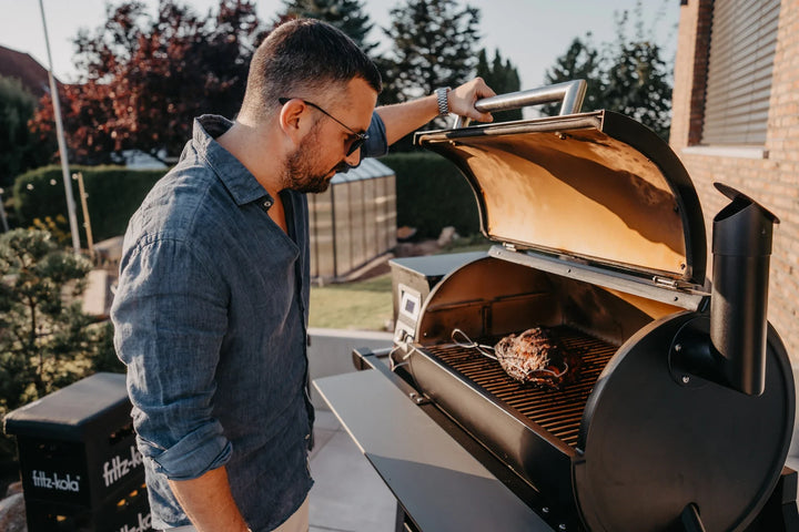 Ein Gast öffnet den MOESTA SHERIFF und schaut auf ein Fleischstück mit Temperaturfühlern auf dem Grillrost.