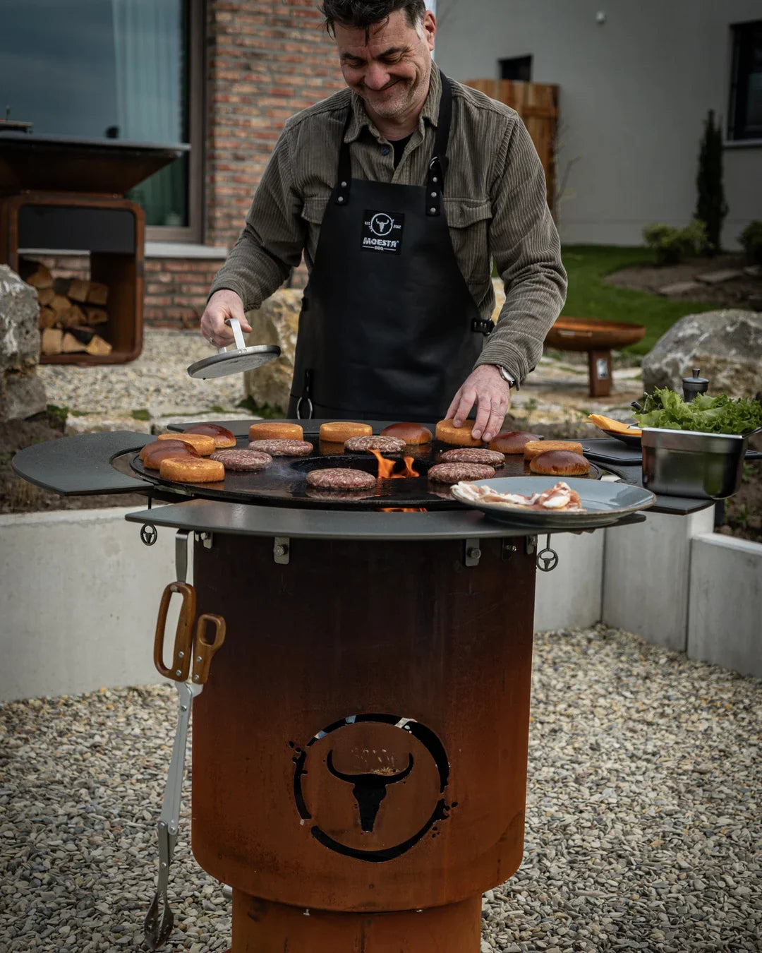 Grillmeister am MOESTA BANDIT 2.0 mit voller BBQ Disk und Burgern über offenem Feuer