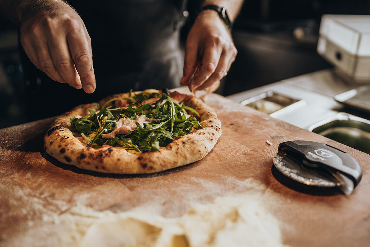 Pizza mit Rucola und Schinken auf einem Holzbrett neben dem PizzaCut