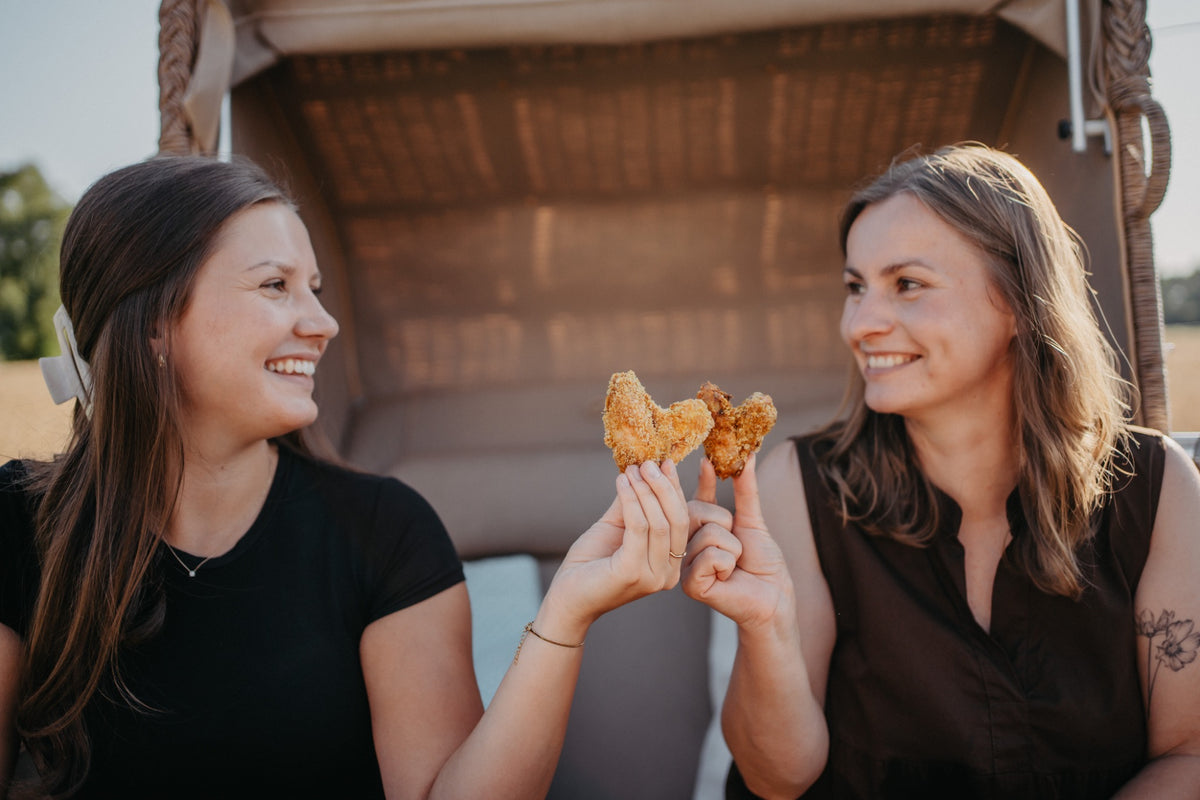 Zwei Freundinnen lachen und halten Chickenwings in Herzform zusammen.