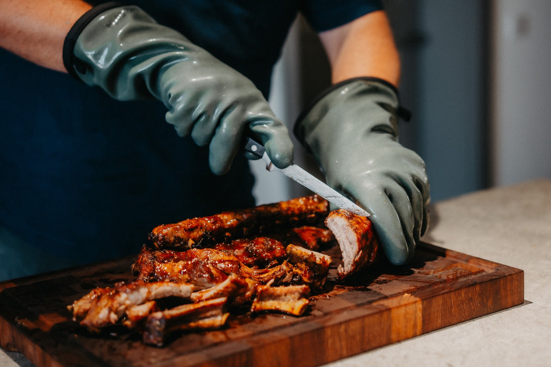 Mit HEATPRO Hitzehandschuhen werden glasierte Spareribs auf einem Holzbrett gehalten und geschnitten.
