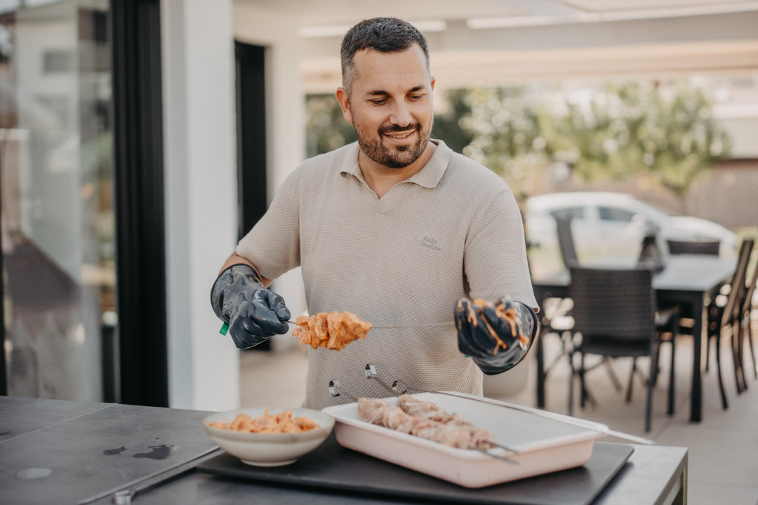 Ein Mann bereitet mit HEATPRO Hitzehandschuhen Grillspieße auf einer hellen Terrasse vor.
